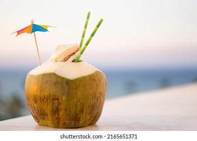 Fresh  Green Coconut Drink With Paper Straw  And Rainbow Umbrella  Standing By The Marble On Tropical Beach Background With Copy Space . Vacation  Exotic  Travel Destinations  Concept .