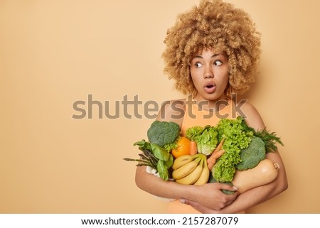 Fresh green food selection. Stunned woman with curly hair embraces green vegetables abd fruits keeps to detox diet has healthy eating poses against beige background blank space for advertisemet