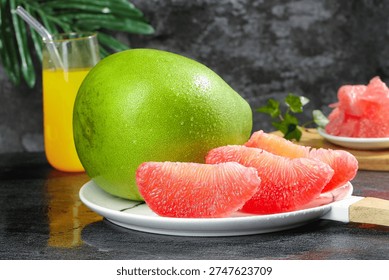 Fresh green pomelo with ruby red grapefruit segments on white plate, juice glass, and tropical fruits displayed on dark surface with green foliage.