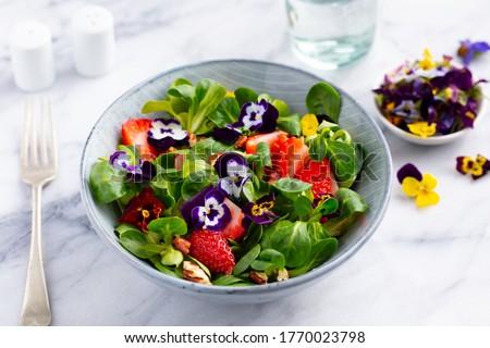 Fresh green salad with strawberries and edible flowers in a bowl. Marble background. Close up.