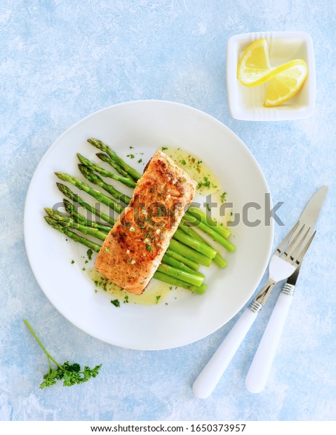 Fresh grilled wild coho salmon\
filet with lemon butter sauce on a bed of steamed organic\
asparagus. White dishes on pale blue background. Healthy eating\
concept.