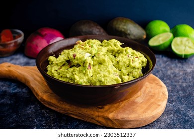 Fresh Guacamole in a Wooden Bowl with Ingredients in the Background: Homemade guacamole with avocados, limes, onion, cilantro, and chipotle peppers - Powered by Shutterstock