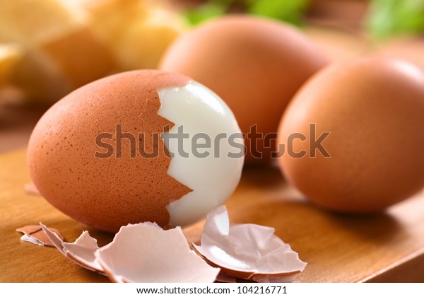 Fresh hard boiled eggs with shell beside on wooden\
board (Selective Focus, Focus on the front of the shell on the\
first egg)
