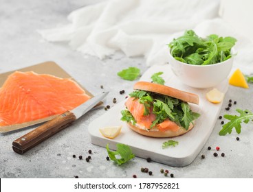 Fresh Healthy Organic Sandwich With Bagel And Salmon, Cream Cheese And Wild Rocket On Chopping Board With Smoked Salmon Pack On Light.