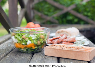 Fresh Healthy Salad With Olives And Feta Chees At Big Bowl On Wooden Table. Baked Cookies, Bread With Sausages At The Picnic Time. Tasty Fresh Food