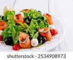 Fresh and healthy salad with smoked salmon, mozzarella, cherry tomatoes, olives and lettuce is served on a white plate