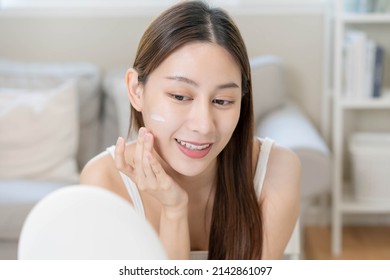 Fresh healthy skin, beautiful smile of asian young woman, girl looking at mirror, applying moisturizer on her face, putting cream treatment before makeup cosmetic routine at home. Facial Beauty.