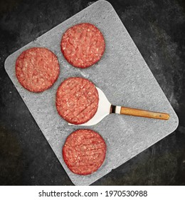 Fresh Home Made Beef Steak Burgers And Spatula On Finnish Fire Stone Slate For Grilling And Serving Hot Dishes. Raw Steak Burgers Cutlets And Grill Tools Overhead View. Burgers For BBQ Grill, Top View