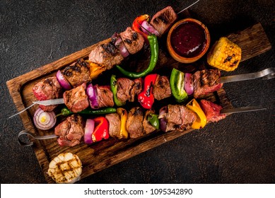 Fresh, home-cooked on the grill fire meat beef shish kebab with vegetables and spices, with barbecue sauce and ketchup, on a dark background on a wooden cutting board above copy space