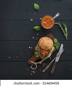 Fresh homemade burger on dark serving board with spicy tomato sauce, sea salt and herbs over dark wooden background. Top view, copy space