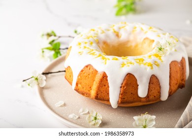 Fresh homemade lemon cake decorated with white glaze and zest on white marble background with branches of blossoming plum.  Close-up.