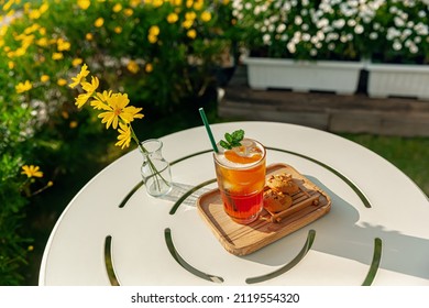 Fresh Homemade Peach Ice Tea With Mint, Chocolate Chip Cookie Served On Table Outdoors. Summer Cold Fruit Drink In Sunny Afternoon With Yellow Flowers Behind