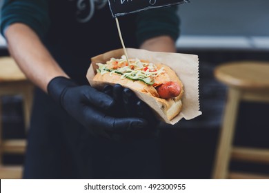 Fresh hot dog with grilled sausage, served outdoors in craft paper. Cookout american bbq food. Hot-dog with meat and vegetables salad filling closeup in chef's hands. Street food, fast food.