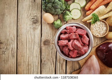 Fresh Ingredients For A Healthy Nutritious Dog Diet With A Bowl Of Chopped Raw Beef, Chicken, Liver, Egg. Assorted Vegetables And Grains In An Overhead View On Wood With Copy Space