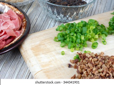 Fresh Ingredients Of A Wild Rice And Brown Rice Dressing Recipe Include Prosciutto, Pecans, Green Onions, And Parsley. The Ingredients Are Scattered On A Wood Table Top And Cutting Board. Narrow Focus