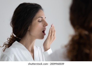 Fresh like wind blow. Millennial hispanic woman wearing bathrobe standing by mirror holding palm by opened mouth checking breath. Young latin female testing breathing smelling air holding palm by lips