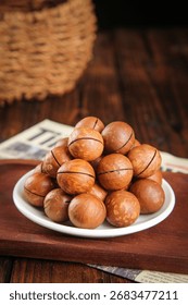 Fresh macadamia nuts with cracked shells arranged on white plate, wooden table background with wicker basket and newspaper, healthy snacking concept