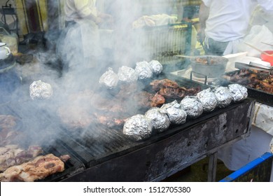 Fresh Meat Pork Or Beef Entricote Grilling On The Grill Outside, Outdoor Setting In The Garden. Picnic Food Festival Event, Warm Ambiance With Steam And Smoke, Cooking Staff And Chef In The Background