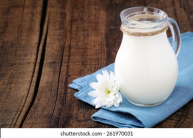 Fresh milk in a glass jug on a blue napkin with a chrysanthemum flower against dark rustic wooden background