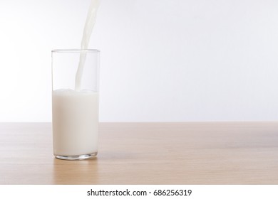 Fresh milk pouring into a half full glass on a plain timber bench top with white background and copy space.