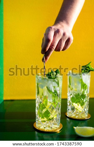 Fresh mojito alcoholic cocktail with rum, lime, mint and ice in a top shot glass shot with hard lights  and a hand stirring it with ice