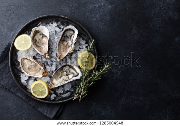 Fresh opened oysters in a plate with\
ice and lemon on black textured background, top\
view