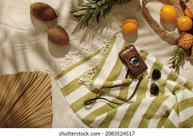 Fresh oranges, pineapple, coconuts falling out from woven bag on sandy beach. Vintage camera, sunglasses on linen beach towel with fringes. Tropical holidays concept