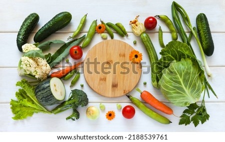 Fresh organic vegetables lie on table around cutting board before preparing vitamin summer meal. Freshly picked: carrot, cucumber, cauliflower, zucchini, tomato, peas, beans, broccoli and cabbage