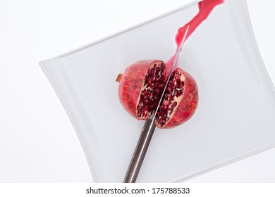 Fresh pomegrate being cut in half with a knife on a white plate with sweet juice running out as the pulpy seeds or arils or exposed, overhead view