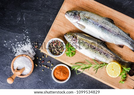Fresh rainbow trouts on wooden board on dark stone table with salt, pepper, lemon and rosemay ingredients. Tasty fishes preparing for lunch.