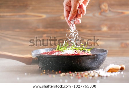 Fresh raw flank steak in the pan. The woman's hand adds salt to the meat. Ingredients are spices, rosemirine various sprouts and finally salt.
