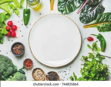 Fresh raw greens, unprocessed vegetables and grains over light grey marble kitchen countertop, wtite plate in center, top view, copy space. Healthy, clean eating, vegan, detox, dieting food concept