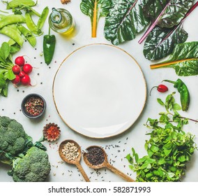 Fresh raw greens, vegetables and grains over light grey marble kitchen countertop, wtite plate in center, top view, copy space. Healthy, clean eating, vegan, detox, dieting food concept