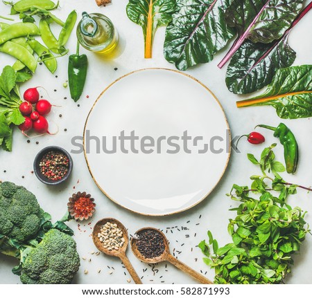 Fresh raw greens, vegetables and grains over light grey marble kitchen countertop, wtite plate in center, top view, copy space. Healthy, clean eating, vegan, detox, dieting food concept