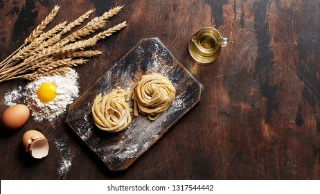 Fresh Raw Homemade Noodles On Vintage Wooden Board With Raw Eggs, Pile Of Flour, Wheat Spikes And Jug Of Olive Oil. Ingredients For Cooking Pasta. Сopy Space, Overhead View 