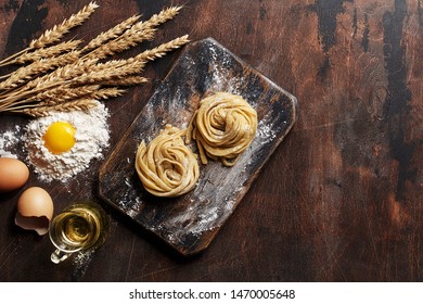 Fresh Raw Homemade Noodles On Vintage Wooden Board With Raw Eggs, Pile Of Flour, Wheat Spikes And Jug Of Olive Oil. Ingredients For Cooking Pasta. Overhead View, Copy Space