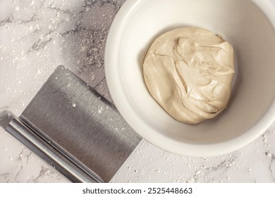 Fresh, raw and homemade pizza dough in a white bowl with a metal scraper on a white marble kitchen counter.  - Powered by Shutterstock