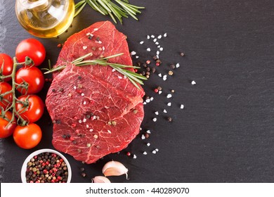 Fresh Raw Meat Beef Steak With With Spices, Rosemary, Tomatoes And Olive Oil On The Black Slate Surface. Top View.