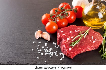 Fresh Raw Meat Beef Steak With With Spices, Rosemary, Tomatoes And Olive Oil On The Black Slate Surface.