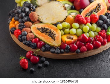 Fresh raw organic summer berries and exotic fruits in round large tray on black kitchen background. Papaya, grapes, nectarine, orange, raspberry, kiwi, strawberry, lychees, cherry. Top view