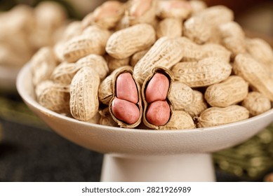 Fresh raw peanuts in shells with red-skinned kernels exposed in white bowl, showcasing natural texture and deep glistening appearance of wholesome nuts