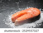 Fresh raw salmon steak with salt on dark textured table, closeup