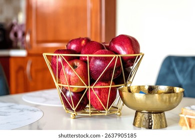 Fresh red apples arranged in geometric golden wire basket placed on kitchen table, next to metallic bowl and marble placemats, healthy lifestyle symbol with warm domestic background.