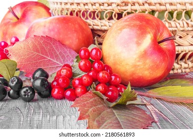 Fresh Ripe Apples, Autumn Leaves And Berries On A Wooden Table Against The Background Of A Wicker Basket With Fruit, Front View, Close-up. Autumn Harvest, Thanksgiving Day Concept. World Vegan Day.