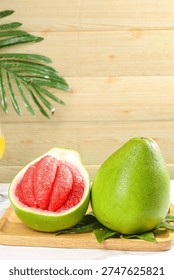 Fresh ruby red grapefruit cut in half displaying vibrant pink flesh alongside whole green grapefruit on wooden surface with tropical palm leaves