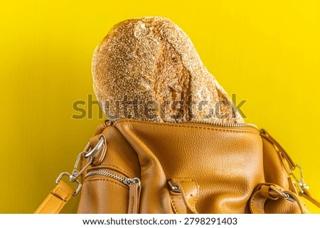 Fresh rustic bread loaf inside a stylish leather handbag on a bright yellow background. Creative concept of grocery shopping, food carrying, daily life, bakery, sustainable lifestyle and unexpected objects.