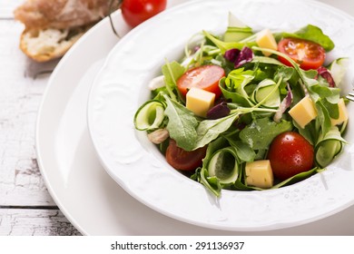 Fresh Salad With Cherry Tomatoes, Arugula, Cucumber And Cheese Cubes In A Plate  Served With Olive Oil And Bread On White Wooden Background. Selective Focus