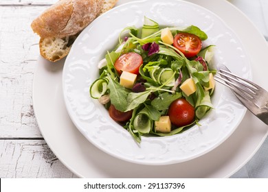 Fresh Salad With Cherry Tomatoes, Arugula, Cucumber And Cheese Cubes In A Plate  Served With Olive Oil And Bread On White Wooden Background. Selective Focus