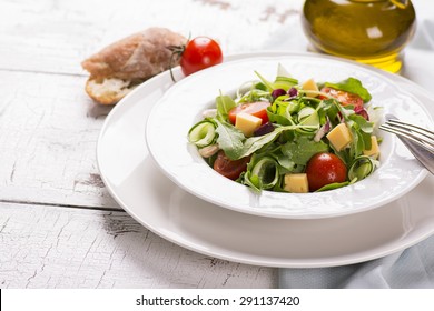 Fresh Salad With Cherry Tomatoes, Arugula, Cucumber And Cheese Cubes In A Plate  Served With Olive Oil And Bread On White Wooden Background. Selective Focus