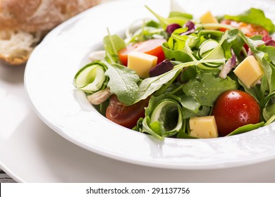 Fresh Salad With Cherry Tomatoes, Arugula, Cucumber And Cheese Cubes In A Plate  Served With Olive Oil And Bread On White Wooden Background. Selective Focus